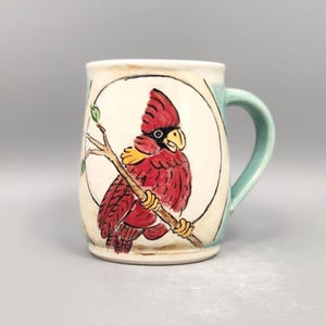May include: Ceramic mug with a hand-painted cardinal bird design. The bird is red with a black head and yellow accents, perched on a branch within a white circle. The mug has a light green handle and rim.