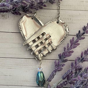 May include: A handcrafted silver heart pendant necklace made from repurposed silverware. The heart is formed from two forks and a woven section. A faceted teal and clear crystal bead hangs below. Lavender sprigs frame the necklace.