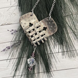May include: A silver-toned metal pendant necklace with a heart-shaped design. The pendant features a woven pattern and is adorned with a cluster of purple beads and a clear crystal. The necklace hangs on a delicate silver chain.