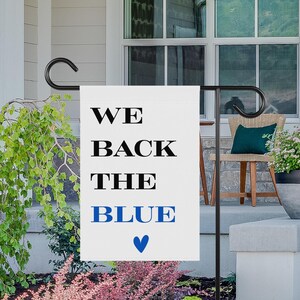 May include: White garden flag with black text that reads "We Back The Blue" with a blue heart at the bottom.