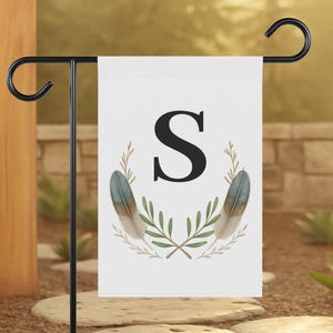 May include: A white garden flag with a black letter "S" in the center, surrounded by a wreath of olive branches and two feathers. The flag is displayed on a black metal stand in a garden setting.