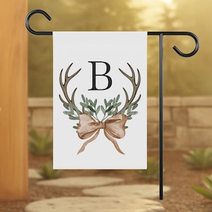 May include: A white garden flag with a black metal stand. The flag features a watercolor design of antlers, greenery, and a bow, with the letter "B" in the center. The flag is displayed outdoors.