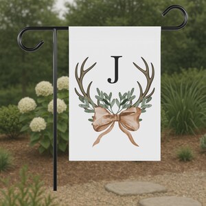May include: A white garden flag featuring a watercolor design with antlers, greenery, and a bow. The letter "J" is centered on the flag. The flag is displayed on a black metal stand in a garden setting.