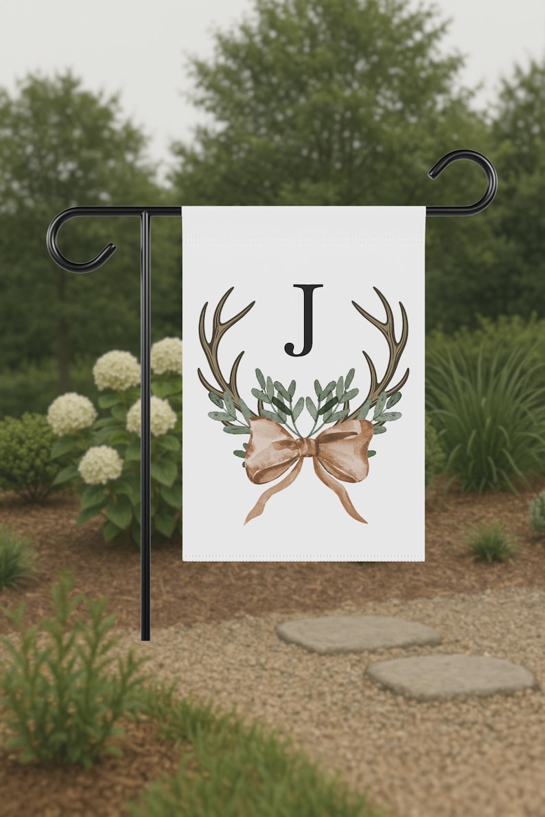 May include: A white garden flag featuring a watercolor design with antlers, greenery, and a bow. The letter "J" is centered on the flag. The flag is displayed on a black metal stand in a garden setting.
