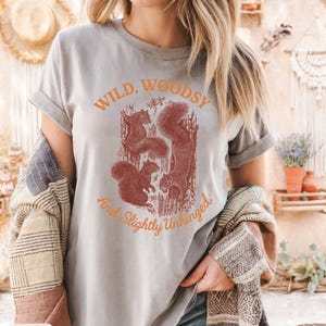 May include: A grey t-shirt with the text "Wild, Woodsy, And Slightly Unhinged" in orange. The shirt features a graphic of squirrels in a tree. The shirt is made of a soft material.