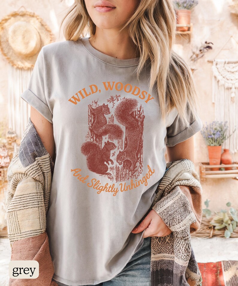May include: A grey t-shirt with the text "Wild, Woodsy, And Slightly Unhinged" in orange. The shirt features a graphic of squirrels in a tree. The shirt is made of a soft material.