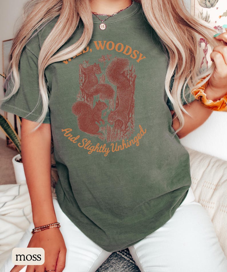 May include: Moss green t-shirt with a squirrel graphic and the text "Wild, Woodsy, And Slightly Unhinged". The shirt features a crew neck and short sleeves. The graphic is in shades of brown and depicts three squirrels in a forest setting.