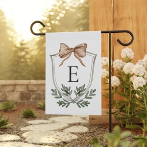 May include: White garden flag featuring a watercolor design with a brown bow, the letter "E", and green leaves. The flag is displayed on a black metal stand in a garden setting.