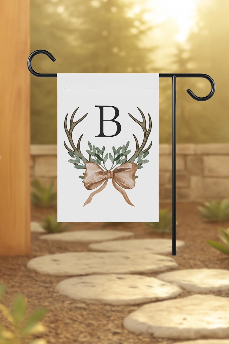 May include: A white garden flag with a black metal stand. The flag features a watercolor design of antlers, greenery, and a bow, with the letter "B" in the center. The flag is displayed outdoors.