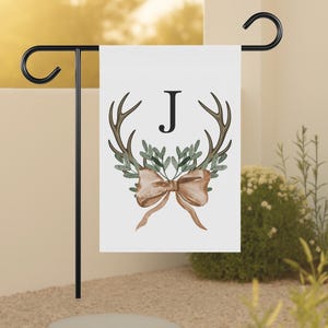 May include: A white garden flag featuring a watercolor design with brown antlers, green leaves, and a tan bow. The letter "J" is centered above the antlers. The flag is displayed on a black metal stand.
