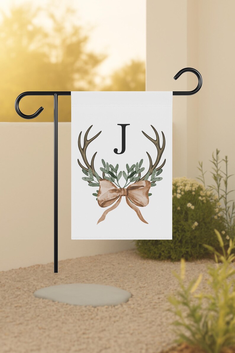 May include: A white garden flag featuring a watercolor design with brown antlers, green leaves, and a tan bow. The letter "J" is centered above the antlers. The flag is displayed on a black metal stand.