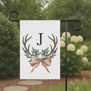 May include: A white garden flag featuring a watercolor design with brown antlers, green leaves, and a tan bow. The letter "J" is centered above the antlers. The flag is displayed on a black metal stand in a garden setting.