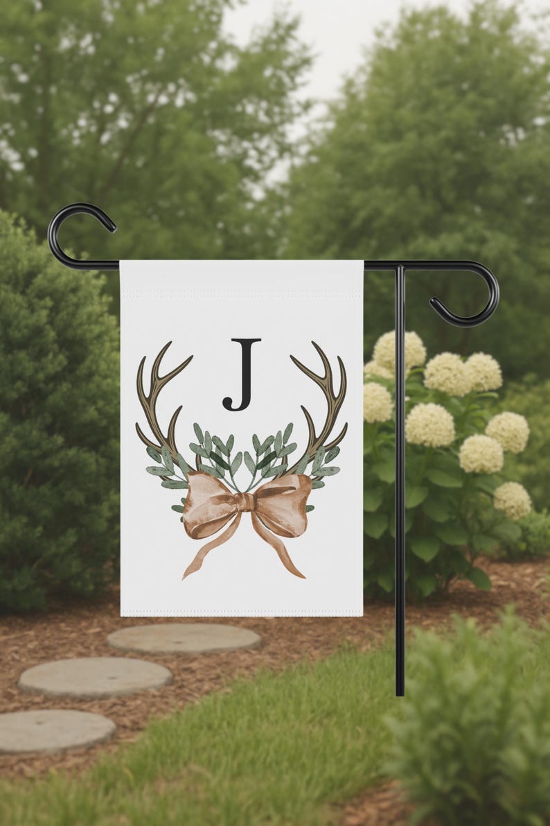 May include: A white garden flag featuring a watercolor design with brown antlers, green leaves, and a tan bow. The letter "J" is centered above the antlers. The flag is displayed on a black metal stand in a garden setting.