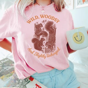 May include: A light pink t-shirt with the words "WILD, WOODSY And Slightly Unhinged" printed in brown. The shirt features a brown graphic of squirrels. A white mug with a yellow smiley face is held in the hand.
