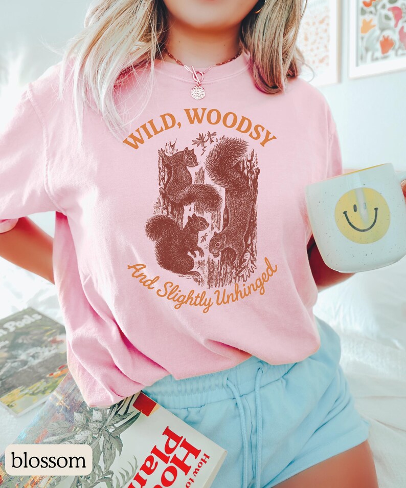May include: A light pink t-shirt with the words "WILD, WOODSY And Slightly Unhinged" printed in brown. The shirt features a brown graphic of squirrels. A white mug with a yellow smiley face is held in the hand.