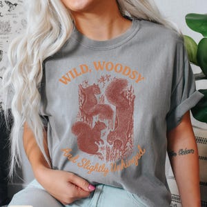 May include: Grey t-shirt with a graphic of squirrels and the text "Wild, Woodsy, And Slightly Unhinged" in orange. The shirt is a relaxed fit.