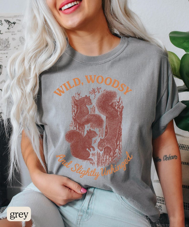 May include: Grey t-shirt with a graphic of squirrels and the text "Wild, Woodsy, And Slightly Unhinged" in orange. The shirt is a relaxed fit.