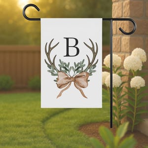 May include: White garden flag with a black metal stand. The flag features a watercolor design of antlers, greenery, a bow, and the letter "B". The flag is displayed outdoors on a sunny day.