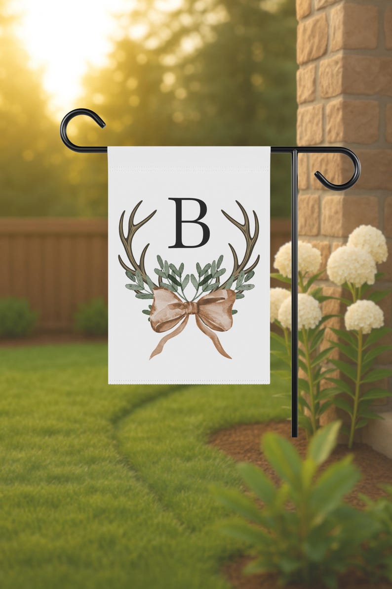 May include: White garden flag with a black metal stand. The flag features a watercolor design of antlers, greenery, a bow, and the letter "B". The flag is displayed outdoors on a sunny day.