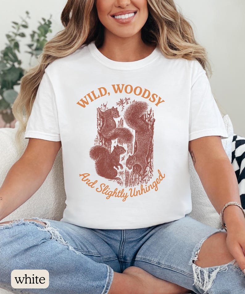 May include: White t-shirt with a crew neck featuring a brown graphic of squirrels and the text "Wild, Woodsy, And Slightly Unhinged" in orange. The shirt is made of a soft material.