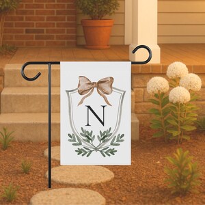 May include: A white garden flag with a black metal stand. The flag features a watercolor design of a bow, the letter "N", and greenery. The flag is displayed outdoors in a garden setting.