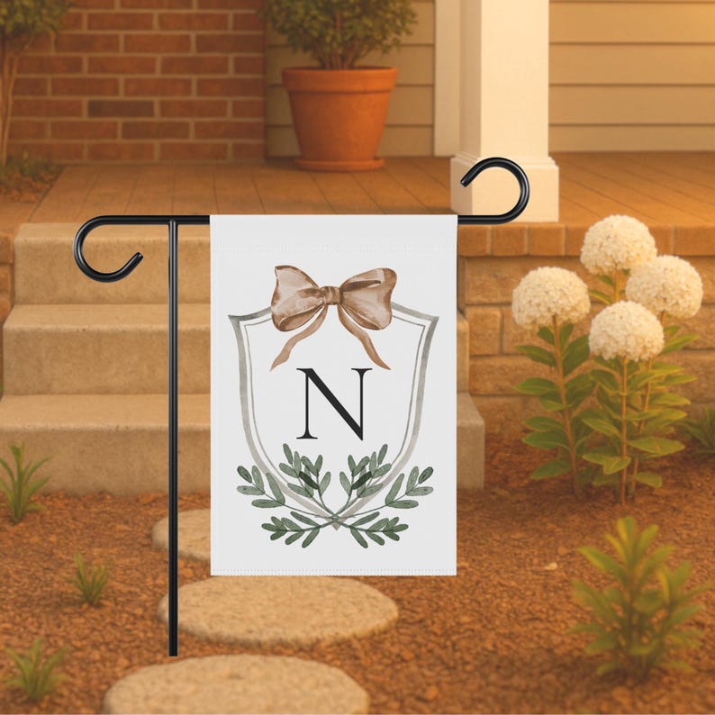 May include: A white garden flag with a black metal stand. The flag features a watercolor design of a bow, the letter "N", and greenery. The flag is displayed outdoors in a garden setting.