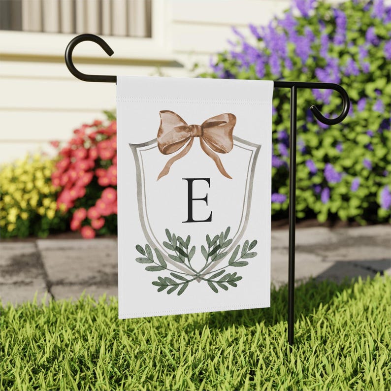 May include: A white garden flag with a brown bow, a gray shield design, the letter "E", and green leaves. The flag is displayed on a black metal stand in a grassy yard with colorful flowers in the background.