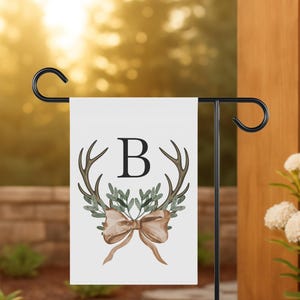May include: A white garden flag with a black metal stand. The flag features a brown antler design, green leaves, a brown bow, and the letter "B". The flag is displayed outdoors.