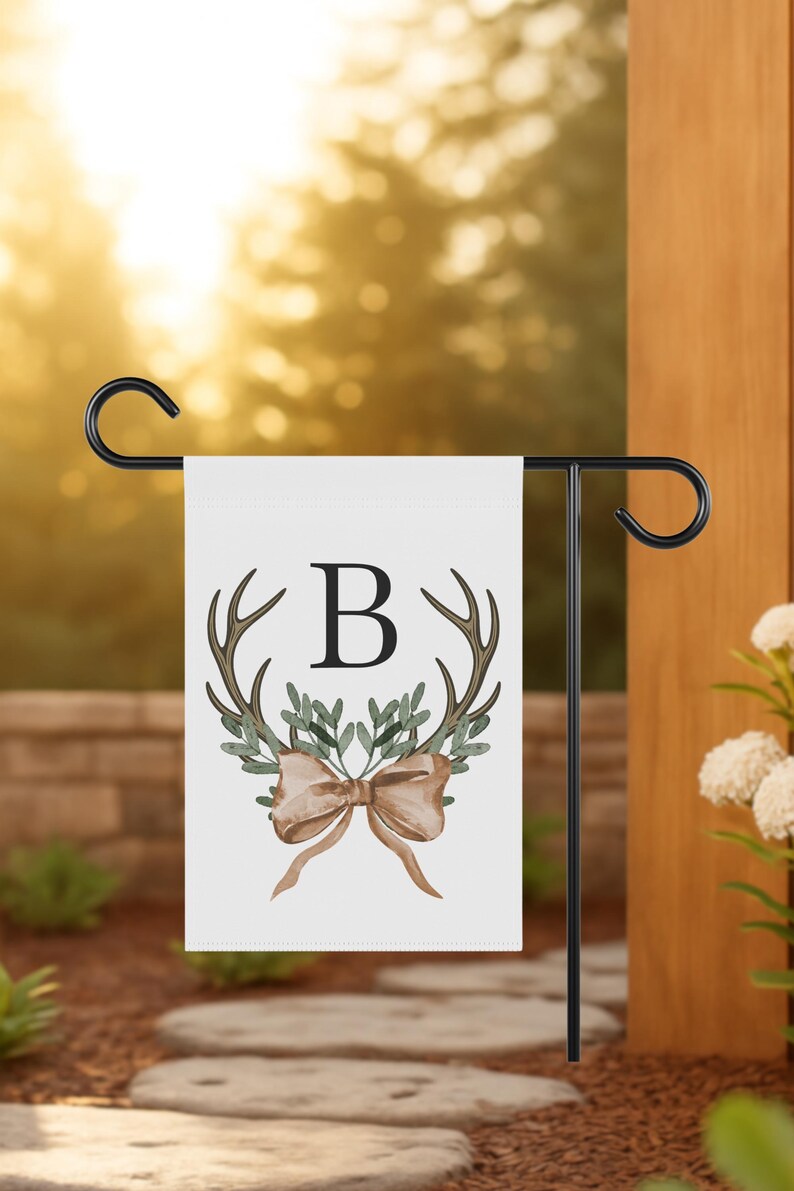 May include: A white garden flag with a black metal stand. The flag features a brown antler design, green leaves, a brown bow, and the letter "B". The flag is displayed outdoors.