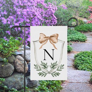 May include: A white garden flag featuring a tan bow, the letter "N", and green foliage. The flag is displayed on a black metal stand in a garden setting with purple flowers and a stone wall.
