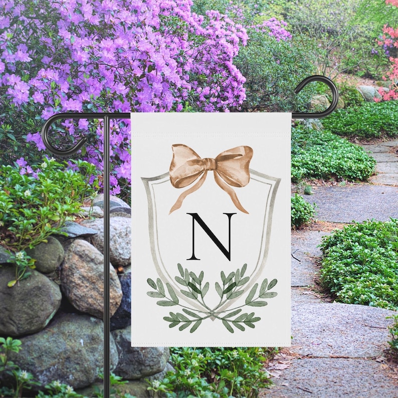 May include: A white garden flag featuring a tan bow, the letter "N", and green foliage. The flag is displayed on a black metal stand in a garden setting with purple flowers and a stone wall.