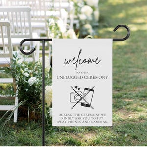 May include: White sign with black text and icons. The sign reads "Welcome to our unplugged ceremony. During the ceremony we kindly ask you to put away phones and cameras." The sign has a crossed-out camera and a phone with hearts above it.