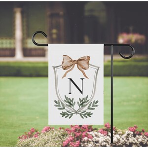 May include: A white garden flag featuring a brown bow, the letter "N", and green foliage. The flag is displayed on a black metal stand, set against a blurred outdoor background with pink flowers.