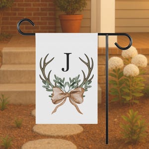 May include: A white garden flag with a black metal stand. The flag features a watercolor design of antlers, greenery, and a bow, with the letter "J" in the center. The flag is displayed outdoors.