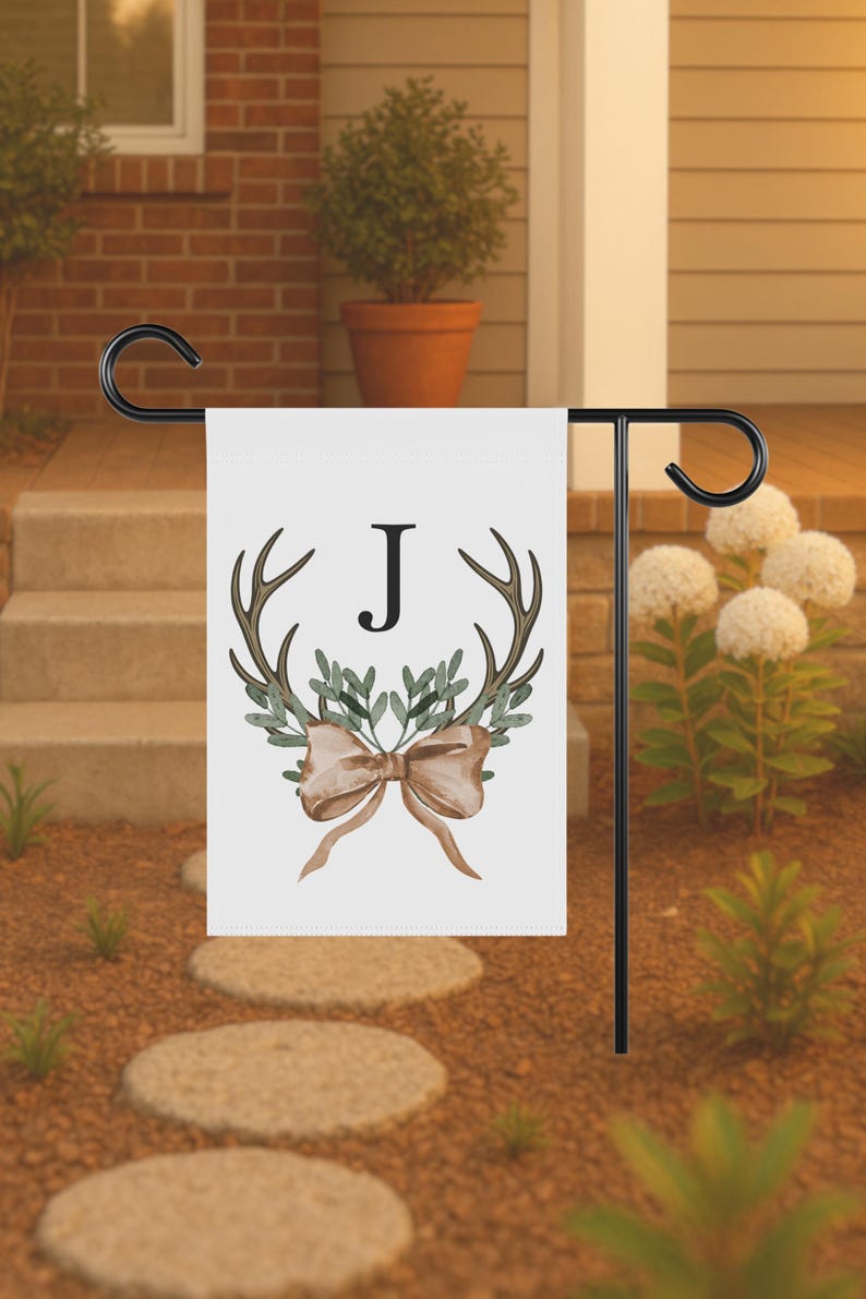 May include: A white garden flag with a black metal stand. The flag features a watercolor design of antlers, greenery, and a bow, with the letter "J" in the center. The flag is displayed outdoors.