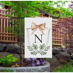 May include: A white garden flag featuring a watercolor design. The flag displays a tan bow, the letter "N", and green foliage on a shield-shaped emblem. The flag is hanging on a black metal stand in a garden setting.