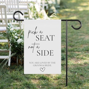 May include: White wedding sign with black text that reads "pick a seat not a side. You are loved by the groom & bride." with a small heart symbol. The sign is attached to a black metal garden stake.