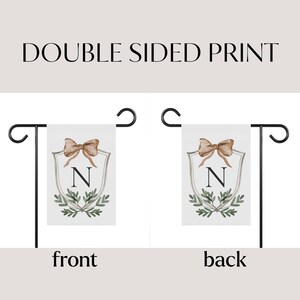 May include: Double-sided garden flag with a white background. The flag features a shield design with a bow, the letter "N", and greenery. The text "DOUBLE SIDED PRINT" is at the top. The front and back of the flag are shown.