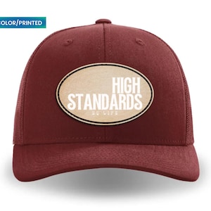 Può includere: Cappellino da camionista bordeaux con retro in rete e visiera curva. La parte anteriore presenta una toppa ovale con le parole "HIGH STANDARDS EQ LIFE" in lettere bianche. Il cappellino ha una chiusura a scatto e un'etichetta colorata/stampata.