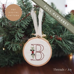 Pode incluir: Um close-up de um ornamento de tecido branco com um design de azevinho bordado em vermelho e verde e a letra "B" em um aro de bordado de madeira. O ornamento está pendurado em uma fita branca em um galho de árvore de Natal verde.