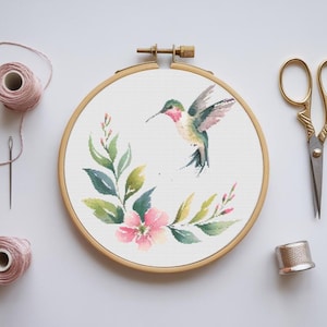 Peut inclure: Un cerceau de broderie au point de croix avec un motif aquarelle d'un colibri avec des fleurs roses et des feuilles vertes.