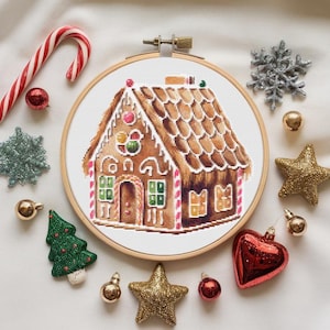 May include: A cross-stitch embroidery hoop with a gingerbread house design. The house is brown with white icing and red candy accents. The hoop is surrounded by various Christmas ornaments, including a candy cane, a snowflake, and a star.