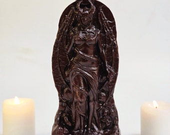 May include: A dark brown statue of Lilith, with horns and wings, standing on a base with the name "LILITH" carved into it. Two white candles flank the statue, casting a warm glow. The statue is intricately detailed.