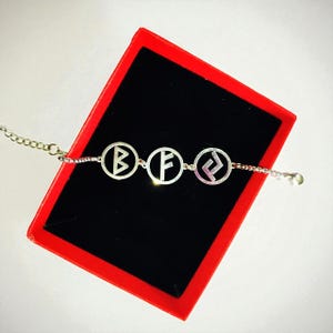 May include: A silver bracelet with three circular charms, each displaying a different rune symbol. The bracelet is set against a black background within a red box, creating a striking contrast. The chain is delicate and the charms are detailed.