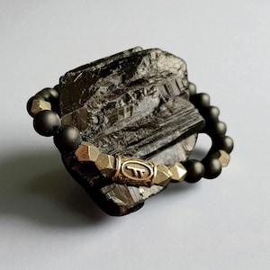 May include: A black beaded bracelet with gold-colored geometric beads and a central gold-colored cylindrical bead with a symbol. The bracelet is resting on a dark, textured stone.
