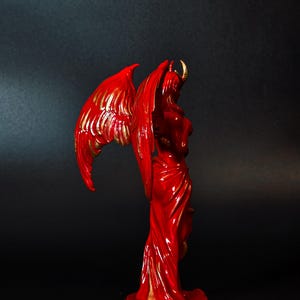 May include: A red and gold figurine of a winged demon with horns. The demon is standing on a base with a gold rim.