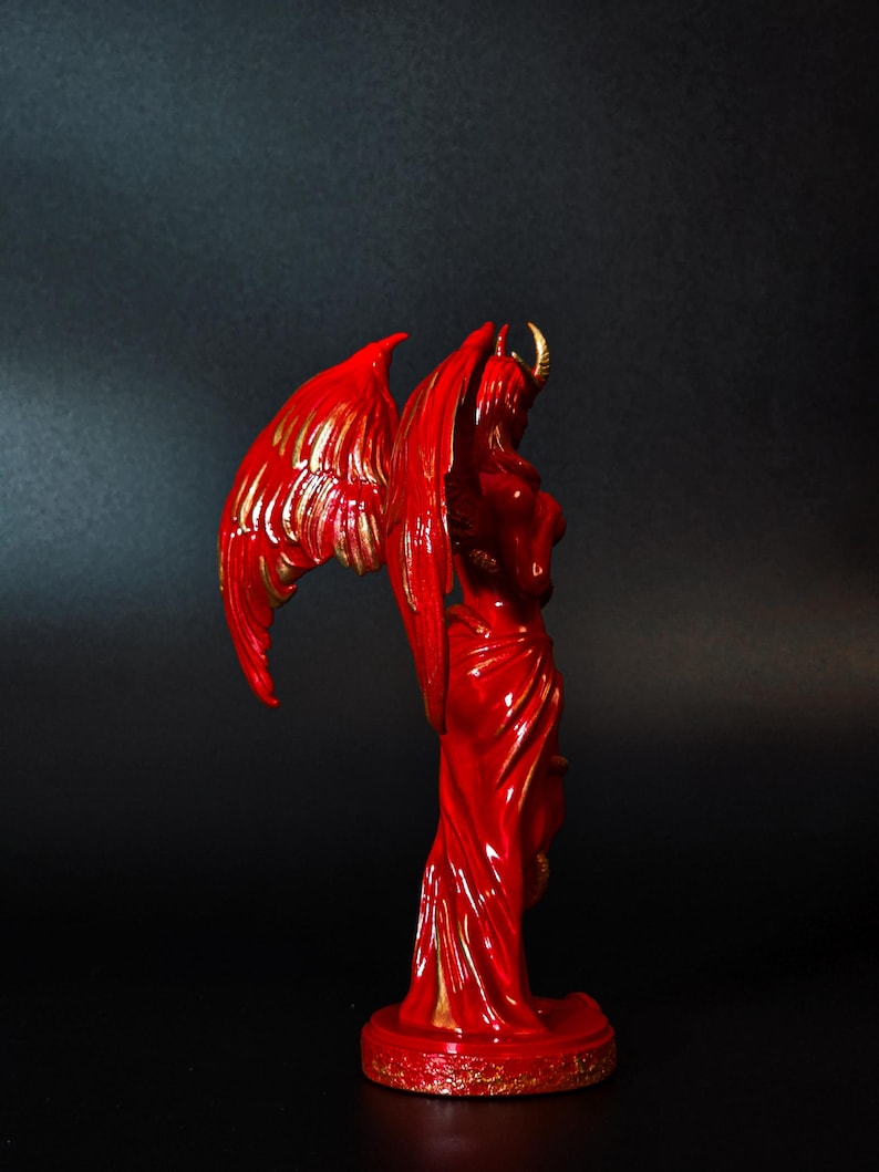 May include: A red and gold figurine of a winged demon with horns. The demon is standing on a base with a gold rim.