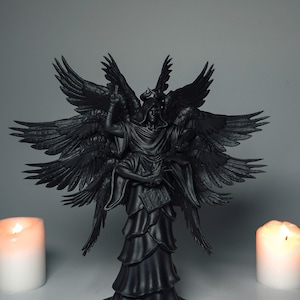 Puede incluir: Una estatua negra de un ángel con múltiples alas, sosteniendo un libro. El ángel lleva una túnica con capucha. Dos velas encendidas flanquean la estatua. La estatua está sobre una base redonda.