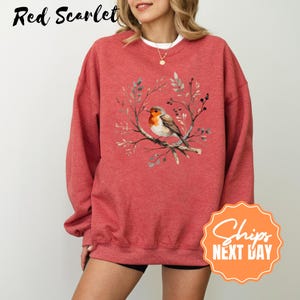 May include: Red scarlet sweatshirt featuring a watercolor illustration of a robin perched on a branch surrounded by leaves and berries. The bird has a red breast and brown wings. The sweatshirt is a solid red color.