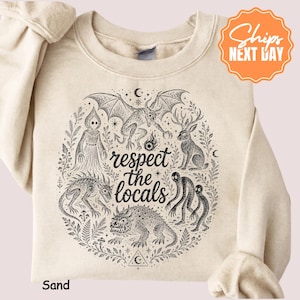 May include: A sand-colored crewneck sweatshirt featuring a black ink-style graphic of various mythical creatures and the text "respect the locals" in the center, surrounded by botanical elements, stars, and crescent moon symbols.